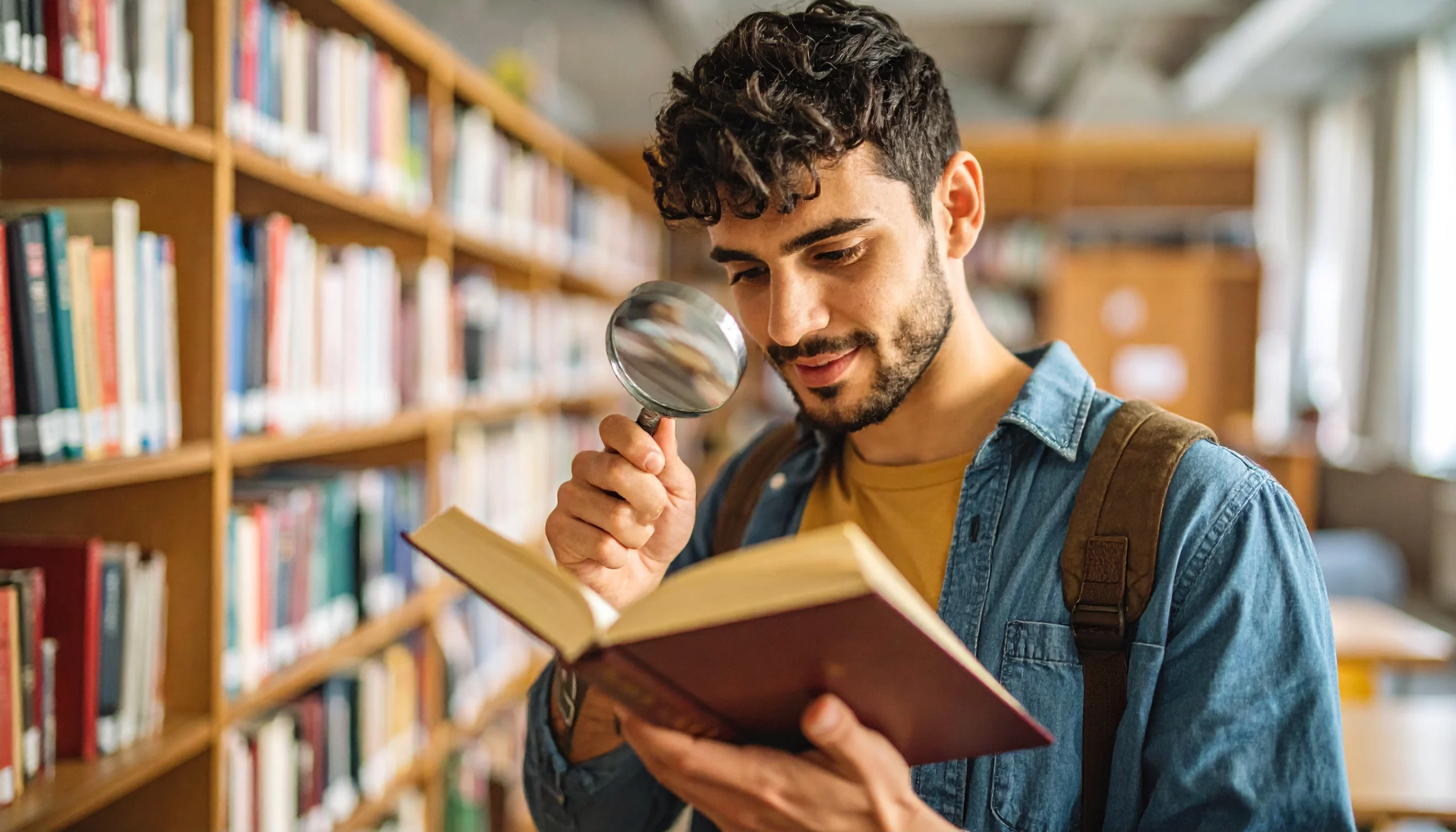 Estudiante universitario en biblioteca revisando un libro con lupa, representando la búsqueda y análisis de los diferentes tipos de objetivos de investigación para tesis, ideal para ilustrar una guía sobre cómo elegir y redactar objetivos exitosos en proyectos académicos.